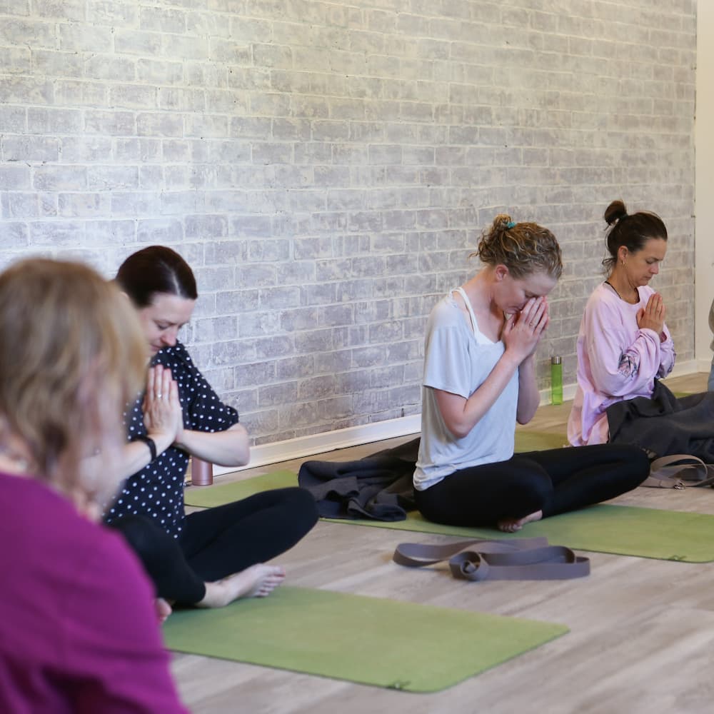 Students seated in a yoga class with hands in prayer position on green mats